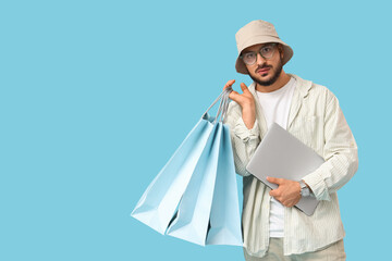 Stylish young man with laptop and shopping bags on blue background
