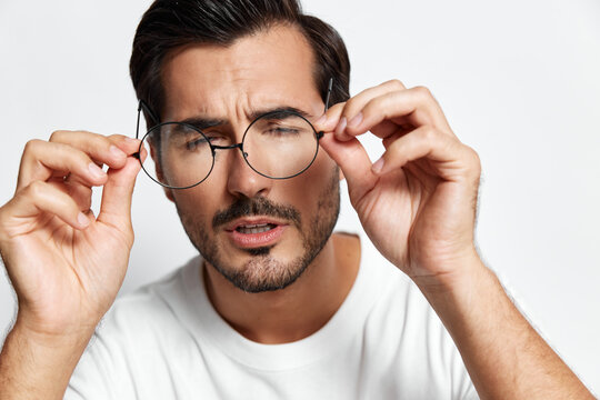 Focused man adjusting his eyeglasses, looking thoughtfully at the camera with a serious expression. Casual style in a simple white t-shirt, studio portrait.