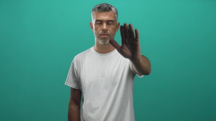 Man middle age with grey hair in white tshirt showing raised palm stop gesture toward camera in studio teal backdrop, frontal pose and steady expression; boundary defiance.