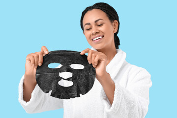 Happy young African-American woman in bathrobe holding activated charcoal mask against blue background