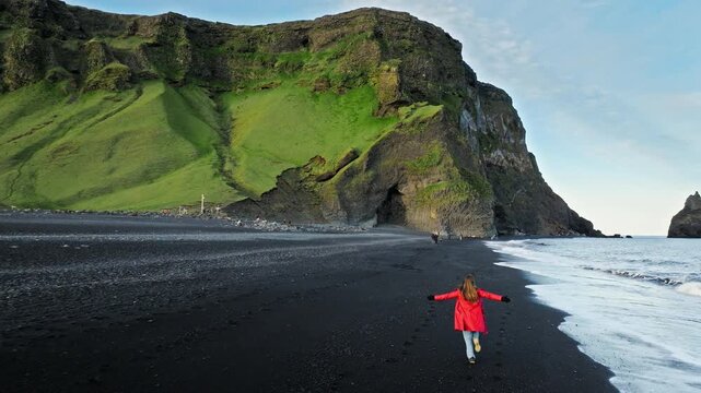 Tourist woman in red coat on Reynisfjara, Iceland, black sand beach. Cinematic 4K drone footage captures Atlantic waves and volcanic cliffs in dramatic scenery.