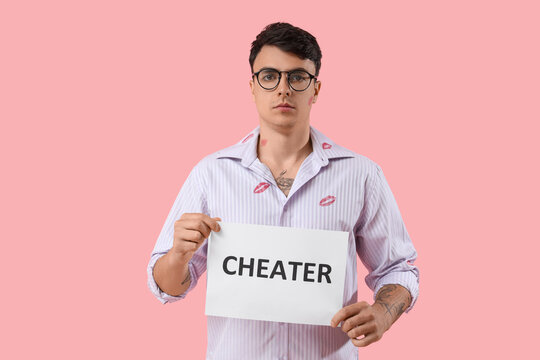 Young man with kiss marks on his shirt holding paper sheet with word CHEATER against pink background