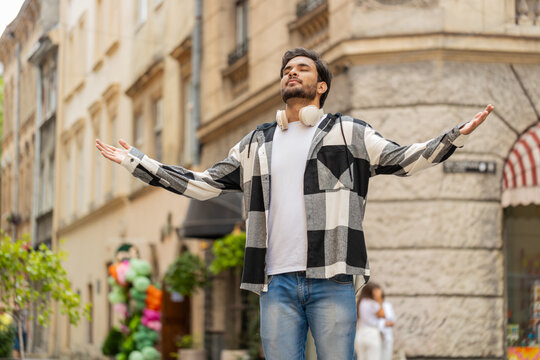 Keep calm down, relax. Young Indian man meditating, breathes deeply with mudra gesture, praying eyes closed, concentrated thoughts, peaceful mind outdoors. Guy taking a break in urban city street.