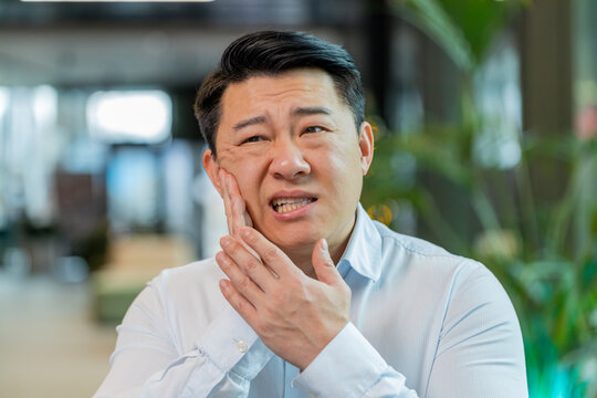 Chinese businessman in office lobby working on laptop and feeling toothache touching cheek with hand, painful expression because of dental illness. Pulpitis, gum disease, dental disease concepts.