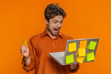 Exhausted freelancer Caucasian man with pasted sticker notes use laptop computer having concentration problem creative crisis. Nervous guy shout confused by big routine work loading orange background