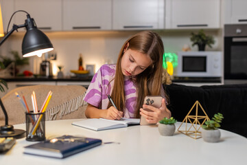 Teenager girl at night home watches online math lesson on smartphone makes notes in notebook. Caucasian child kid sitting at table at evening studies math writes formula solves tasks focus learning.