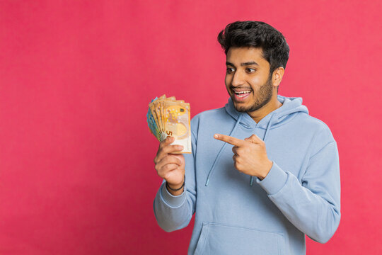 Happy Indian man holding euro cash, counting money with satisfaction, showing profit, salary, or bank loan success. Excited Arabian guy displaying wealth and motivation, isolated on red background