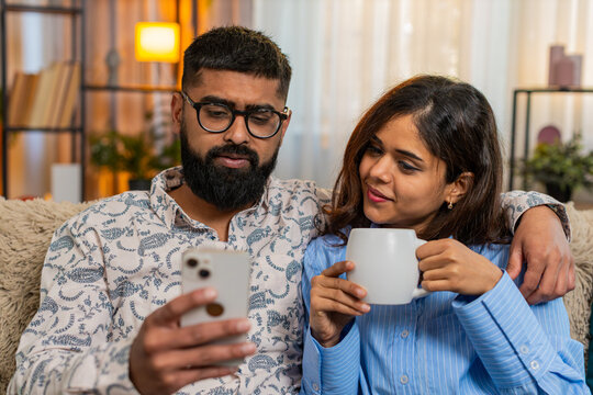 Indian couple man and woman sitting on home sofa, drinking morning coffee and calmly browsing smartphone while talking. Arabian family enjoying peaceful start of day and discussing plans news indoors