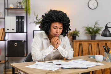 African American businesswoman at home office views bills on invoices feeling stressed about debt and taxes. Freelancer girl at table holds receipts worried by fees budget gap cuts to avoid bankruptcy