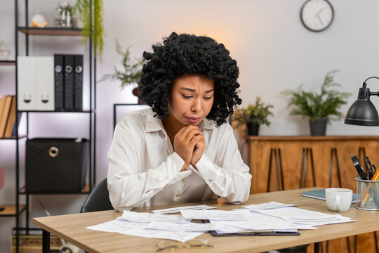 African American businesswoman at home office views bills on invoices feeling stressed about debt and taxes. Freelancer girl at table holds receipts worried by fees budget gap cuts to avoid bankruptcy - Powered by Adobe