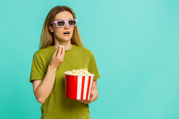 Middle-aged woman wears 3D glasses, holds popcorn watches exciting movie with amazed look, reacting to thrilling surprising storyline. Girl on blue background shows cinematic engagement and emotion