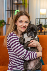 Obraz premium Middle-aged woman hugs French bulldog at home smiles enjoys warm cuddle close bond calm joy. Girl with dog on sofa feels happy relaxed gives care and affection cozy portrait closeup for stock search
