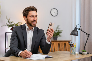 Middle-aged businessman at home office joins smartphone video call listens wearing wireless earphones. Freelancer guy at table writes notebook notes smiling friendly to remote partner.