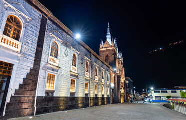 Illuminated Basilica of Our Lady of the Holy Water stands in Banos de Agua Santa, Ecuador. Semi-Gothic church features volcanic stone architecture and streetlights under night sky