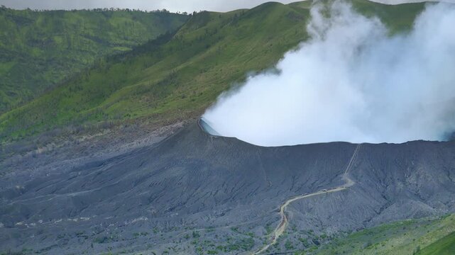Broad green plain and active volcano Mount Bromo emitting white smoke and dust during eruption. Tengger Semeru National Park. East Java, Indonesia. Bird's eye view. Long shot. Camera zooming out