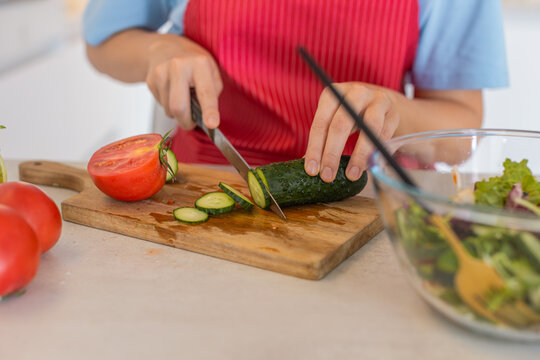 Young woman at home in kitchen cuts cucumber into rounds on wooden board next to chopped tomato. Hands in apron move knife slices into bowl with greens clean counter fresh prep healthy salad making.