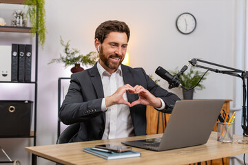 Middle-aged businessman at home office streaming with earphones receives donation shouts wow forms heart gesture. Freelancer guy at table gratefully thanks audience smiling and promising new content.