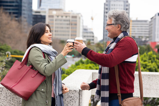 Diverse couple raising coffee cups, leaning on railing on urban terrace with tote and crossbody bag