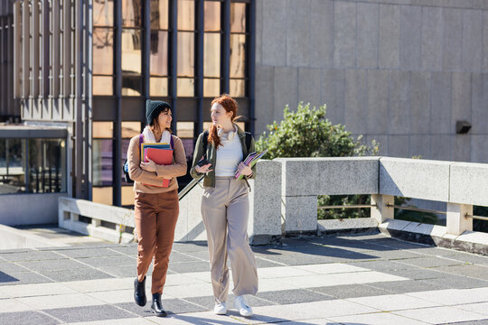 Diverse female students walking along campus path carrying folders notebooks smartphones headphones