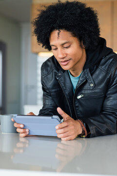 Man leaning on modern home kitchen countertop holding tablet while wearing leather jacket near mug