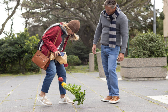 Senior couple bending forward on park walkway spilling celery and tomato from paper grocery bag