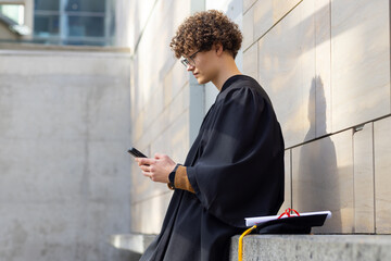 Male graduate in gown sitting on campus bench using smartphone, mortarboard and diploma, copy space