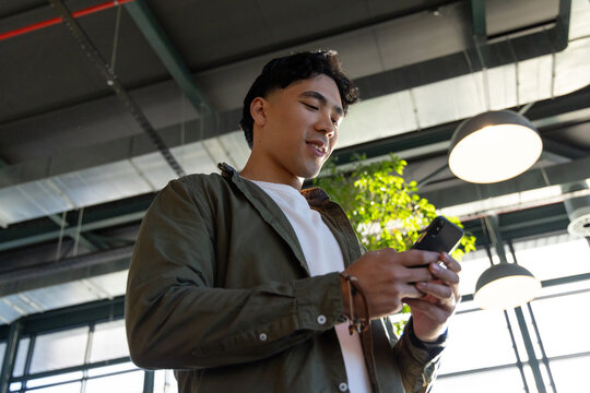 Asian man holding smartphone reading screen in office with leather bracelets, plant, pendant lamps