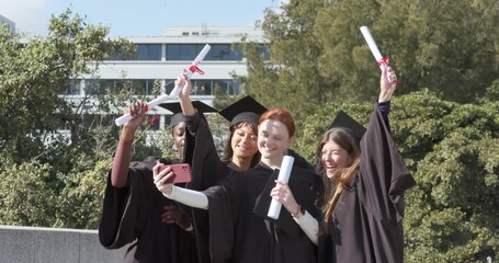 Diverse female graduates in gowns mortarboards snapping selfie with diplomas pink phone on terrace - Powered by Adobe