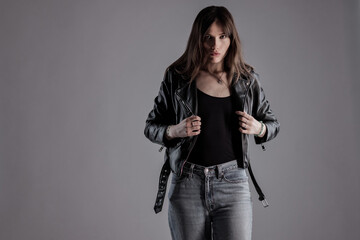 Fototapeta premium Studio fashion portrait of a young woman wearing a black leather jacket and jeans on a gray background. This image is suitable for beauty style and fashion concepts.