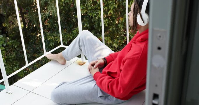 Camera panning red hooded adult placing headphones on balcony, gazing at green foliage listening