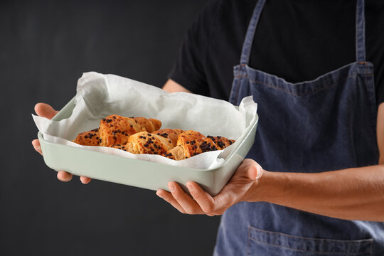Handsome young man with baking dish of fresh croissants on black background - Powered by Adobe