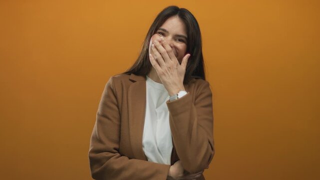 Hispanic woman laughing warmly against an orange wall, covering her mouth with her hand, wearing a brown jacket and a white shirt, with long hair and a watch in view.
