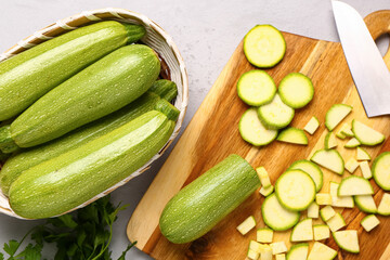 Wicker bowl with many fresh green zucchini and board of slices on grey background