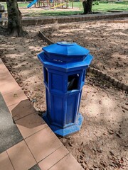 Naklejka premium Octagonal blue trash bin in public park with playground, tiled walkway, and scattered dry leaves