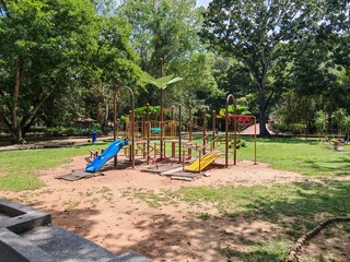 Playground with ramps, slides, and climbing structures in shaded green park with grass and trees