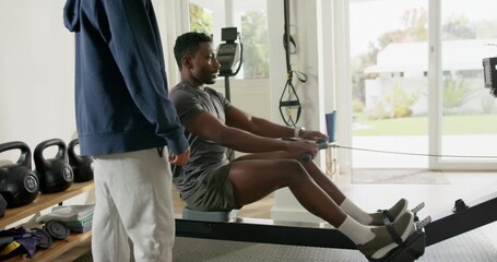 Trainer cueing mid adult African American man rowing on rowing machine for strength in home gym - Powered by Adobe