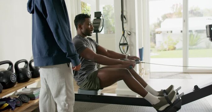 Trainer cueing mid adult African American man rowing on rowing machine for strength in home gym