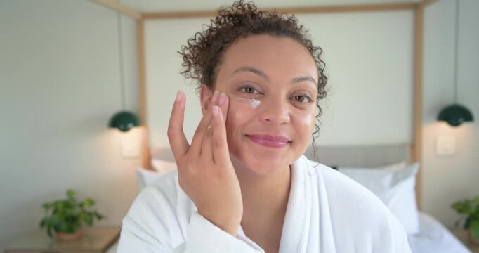 African American woman looking at camera dabbing cream under eyes in bedroom wearing robe showing