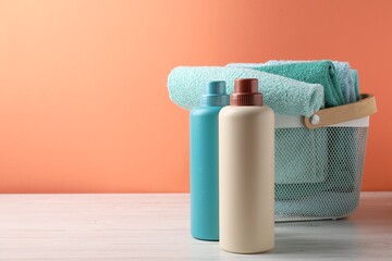Laundry basket with towels and bottles of detergent on white wooden table against coral background, space for text