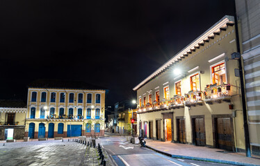 Colonial architecture lines San Francisco Square in historic center of Quito, Ecuador. White building features balconies with flower pots illuminated by streetlights under night sky