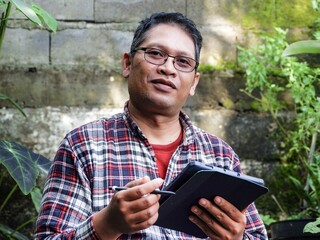 Smiling mature Asian man in a plaid shirt wearing glasses and holding a stylus with a tablet. A creator finding fresh ideas while working in a bright, green backyard garden.