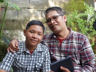 Portrait of a smiling Asian father in a plaid shirt with his arm around his teenage son in a green garden. Concept of family bonding, love, and happiness in nature.