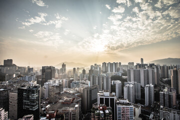 A dramatic cityscape with high-rise buildings under a partly cloudy sky, sun rays piercing through, and distant mountains framing the horizon.