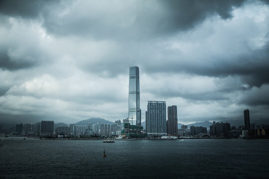 A dramatic view of Hong Kong&rsquo;s skyline with the ICC tower under dark clouds, overlooking Victoria Harbour and distant mountains.
