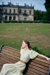 Fototapeta premium Woman in a pale vintage dress sits on a wooden bench in a sunny park, with a historic building in the background, radiating calm and timeless elegance.