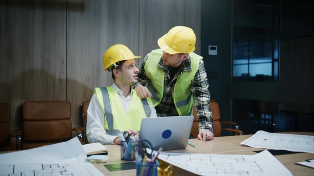 Two male engineers discuss project blueprints and plans in an office during work hours with safety gear on and a laptop open