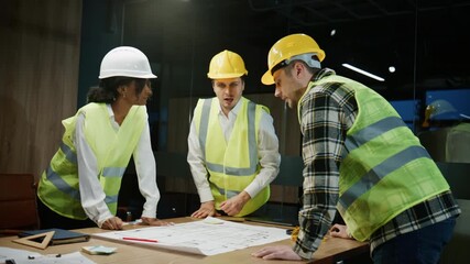 Discussion between multiracial architects and engineers over blueprints in an office space with safety gear