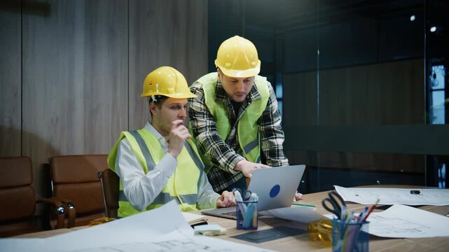 Two architects review project blueprints together in an office setting during working hours in front of a laptop and scattered papers