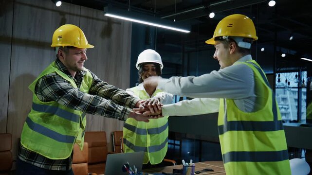 Three multi-racial engineers in protective gear clasp their hands together in an office dedicated to project development, demonstrating teamwork and preparation for the tasks ahead.