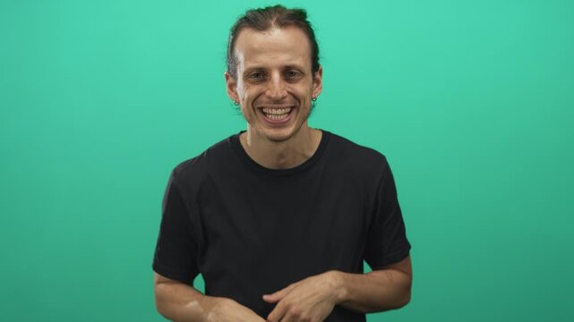 Young hispanic man with long hair sticking tongue and framing face with hands in green studio set; playful mischief energy.